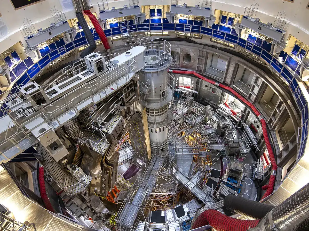 Vedere aeriană fisheye a interiorului halei tokamak ITER în etapa de asamblare, cu platforma circulară, structuri metalice, eșafodaj și conducte de ventilație roșii — șantierul de construcție al celui mai mare reactor de fuziune nucleară din lume